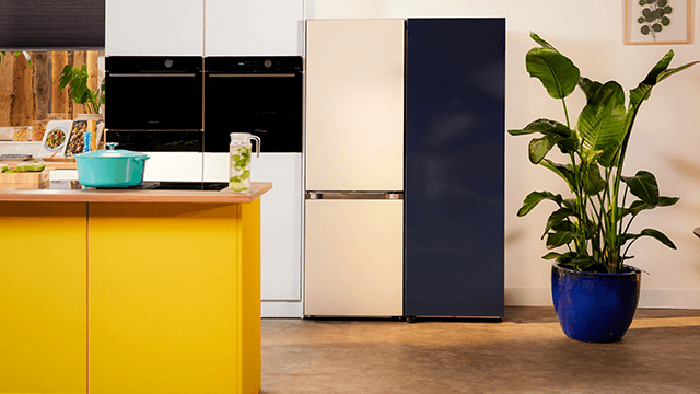 Een gele keuken met een blauwe pan, een koelkast met een beige en blauwe deur, en een plant in een blauwe pot.