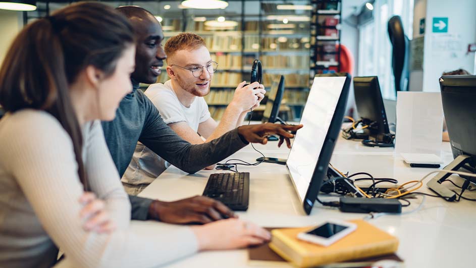 Drie mensen zitten aan een bureau met computers. Een persoon wijst naar het scherm. Boeken en een telefoon liggen op tafel.