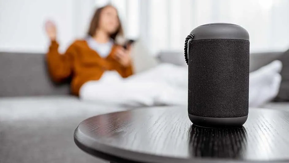 Een zwarte luidspreker staat op een donkere tafel. Op de achtergrond ligt een persoon op een grijze bank met een witte deken.