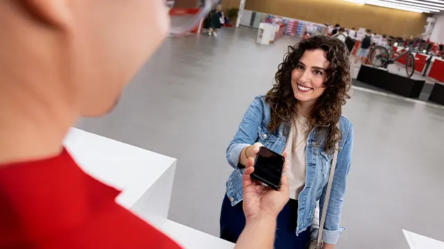 Persoon met krullend haar in spijkerjasje toont smartphone in een winkelruimte met stands op de achtergrond.