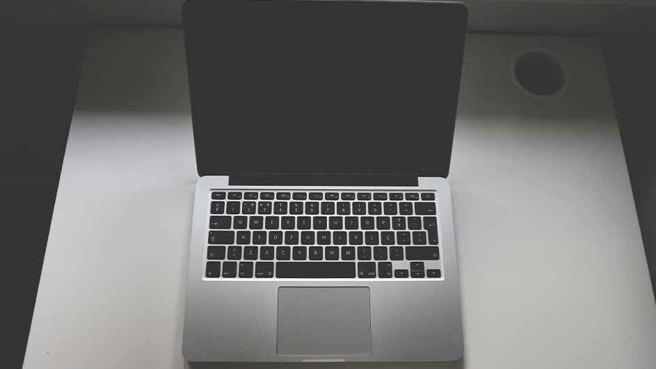 Een opengeklapte laptop staat op een wit bureau. Het scherm is zwart en het toetsenbord is zilverkleurig met zwarte toetsen.