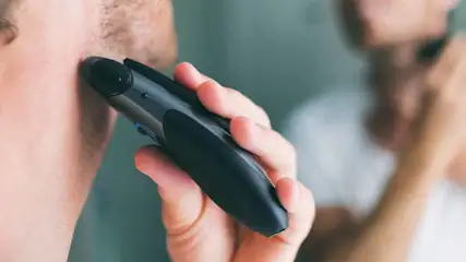 Een man trimt zijn gezichtshaar met een zwarte trimmer. Op de achtergrond staat een persoon voor een spiegel.