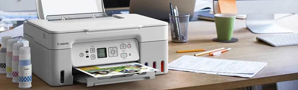 Een Canon‑printer op een druk bureau print in kleur; inktflesjes, notities, potloden, een mok en toetsenbord liggen erbij.