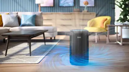 Een zwarte luchtreiniger staat midden in een moderne woonkamer en verspreidt zichtbaar frisse luchtstromen over de houten vloer en de bank.
