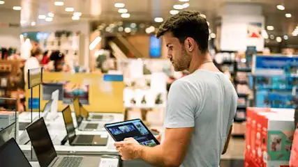 Een man in een winkel bekijkt een tablet, omringd door laptops op een tafel.