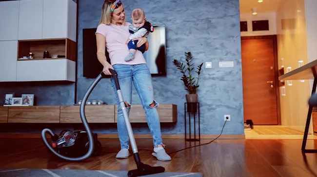 Een vrouw stofzuigt een houten vloer terwijl ze een baby op haar arm houdt, in een moderne woonkamer. Het merk van de stofzuiger is niet zichtbaar.