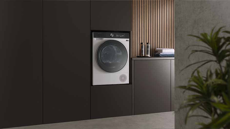 Wasmachine ingebouwd in donkerbruine kasten naast een aanrecht met flessen en handdoeken, houten lambrisering op de achtergrond.