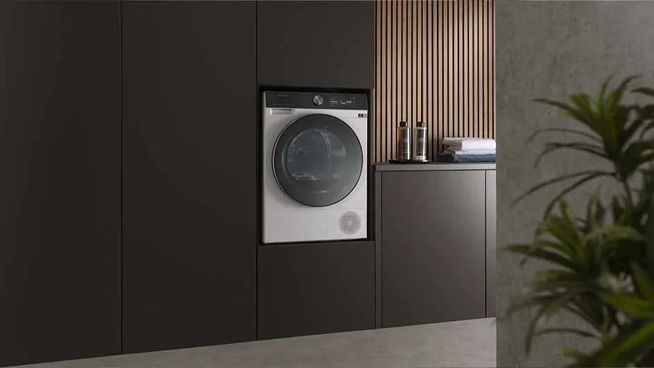 Wasmachine ingebouwd in donkerbruine kasten naast een aanrecht met flessen en handdoeken, houten lambrisering op de achtergrond.