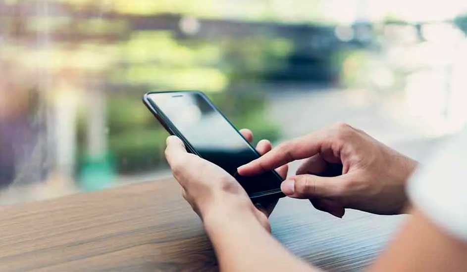 Een persoon bedient een zwarte smartphone met één hand aan een houten tafel bij het raam. Merk van de telefoon is niet zichtbaar.