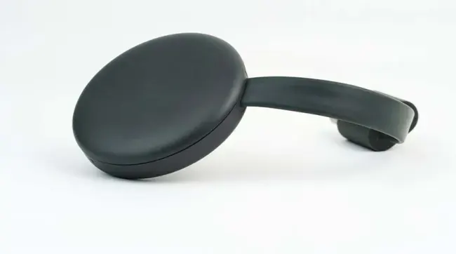 Een donkergrijze Chromecast ligt op een witte ondergrond, met een ronde vorm en een korte, platte kabel.