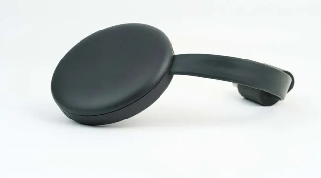 A dark gray Google Chromecast device is on a white surface. The device has a round shape with an attached HDMI connector.