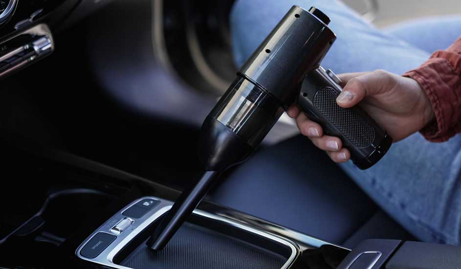 Een persoon gebruikt een compacte zwarte handstofzuiger om het interieur van een auto schoon te maken, vooral bij de middenconsole. Merk: onbekend.