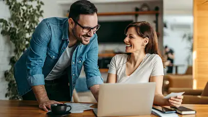 Man met bril en vrouw kijken samen naar laptop, lachen gelukkig, moderne keuken achtergrond, koffiekop en boeken op tafel