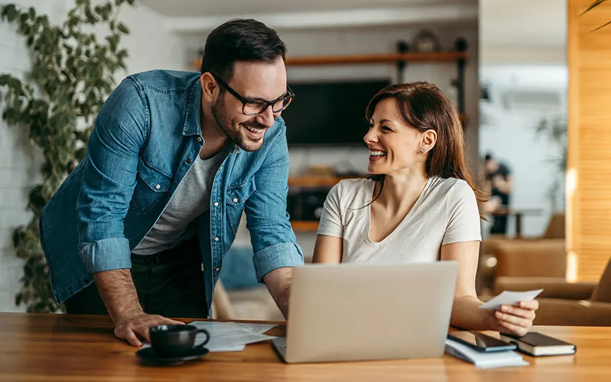 Man met bril en vrouw kijken samen naar laptop, lachen gelukkig, moderne keuken achtergrond, koffiekop en boeken op tafel