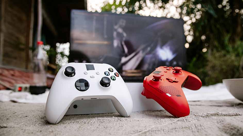 Witte en rode gamecontrollers voor een scherm met een actiespel, op een tafel met een buitenscène op de achtergrond.