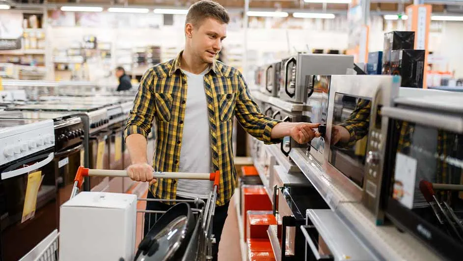 Een man met een winkelwagen bekijkt magnetrons in een winkel. Rekken met apparaten op de achtergrond.