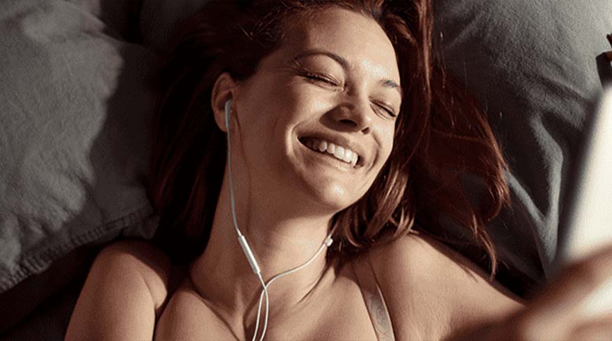 Een lachende vrouw ligt op een grijs kussen met oortjes in en kijkt naar een witte telefoon.