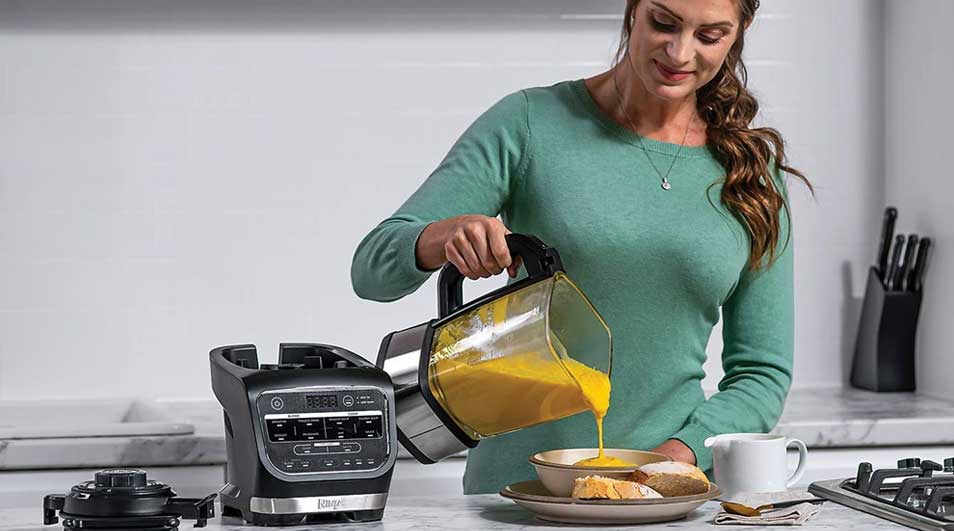 A woman in a kitchen pours yellow soup from a blender jug into a bowl; on the counter sit the blender base, bread and a cup.