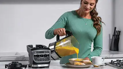 A woman in a kitchen pours yellow soup from a blender jug into a bowl; on the counter sit the blender base, bread and a cup.