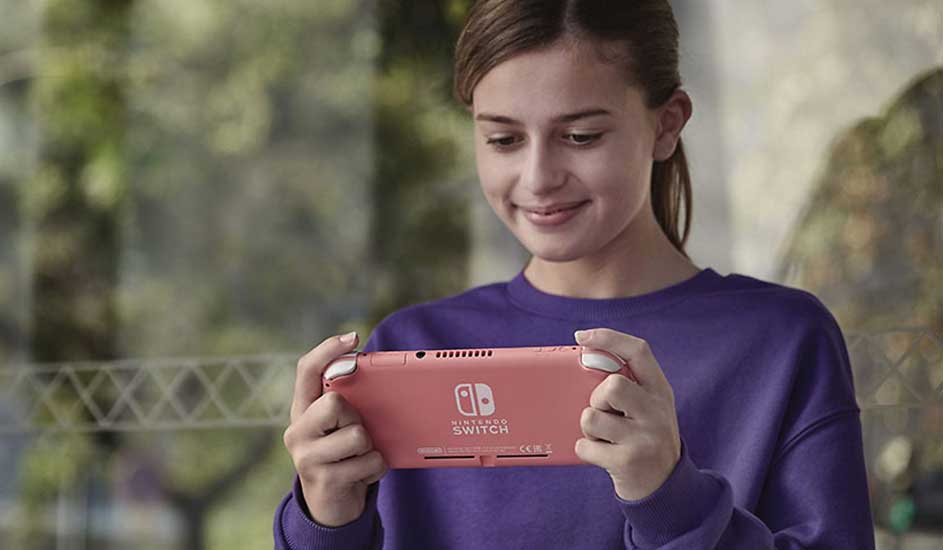 Een persoon in een paarse trui houdt een roze Nintendo Switch Lite vast en speelt, met een groene tuin op de achtergrond.
