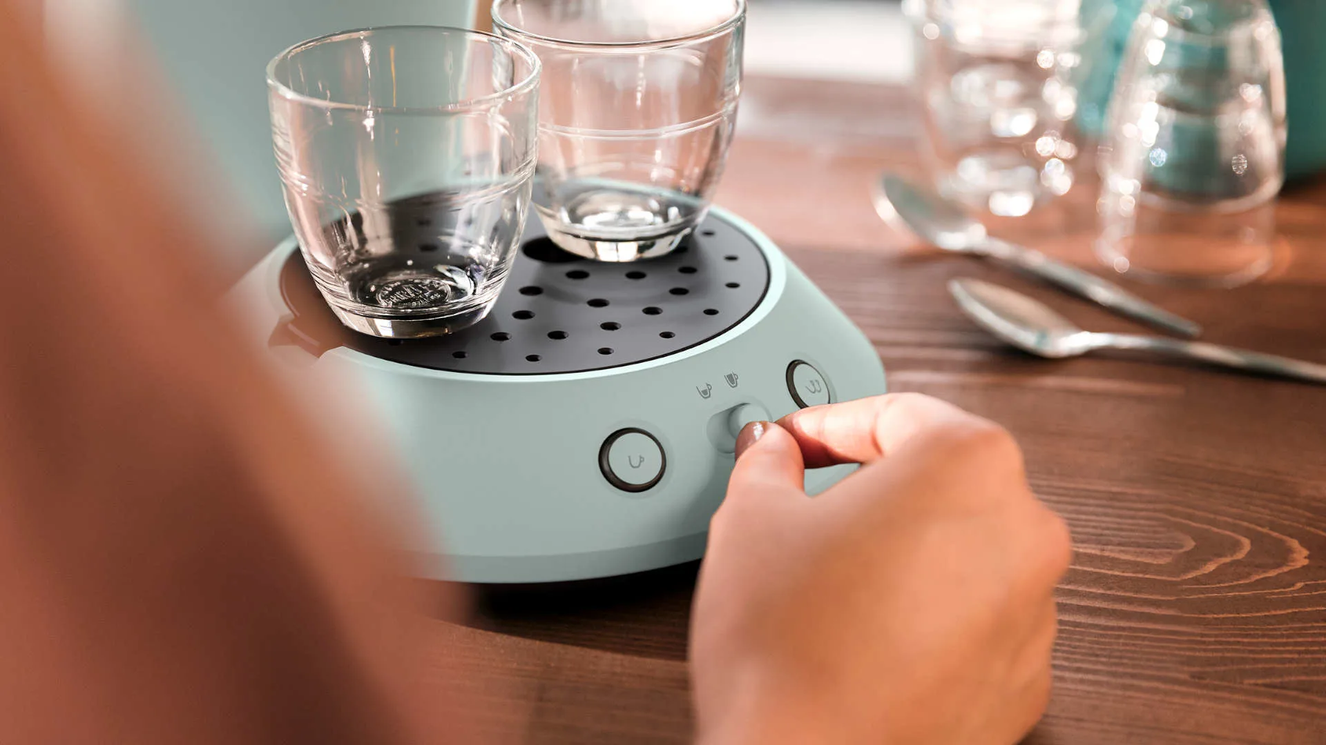 Twee glazen op een turquoise apparaat. Een hand draait aan een knop. Lepels en een glas op de houten tafel.