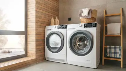 Wasmachines en droger staan naast elkaar in een kamer met een houten ladder en een mand met wasgoed erop.