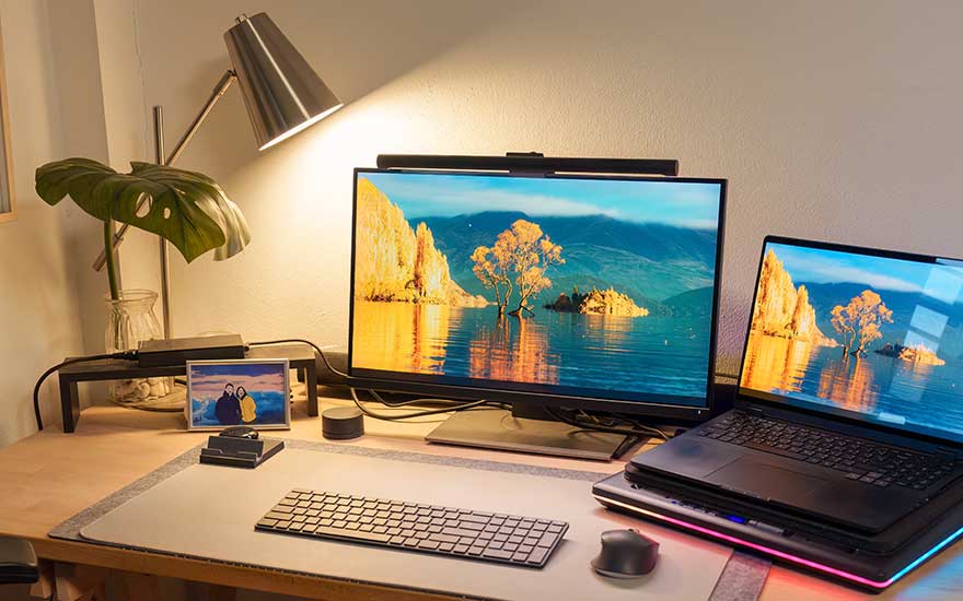 Een bureau met twee monitoren, een foto, lamp en planten in een warme sfeer.