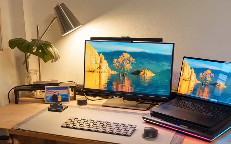 Een bureau met twee monitoren, een foto, lamp en planten in een warme sfeer.