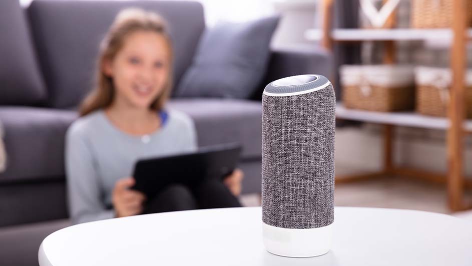 Meisje met tablet op achtergrond, luidspreker op witte tafel. Grijze bank en houten rek in de kamer.