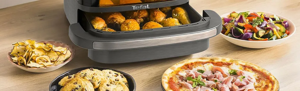 A Tefal air fryer sits on the table with chicken, biscuits, pizza, chips and a bowl of grilled vegetables around it.