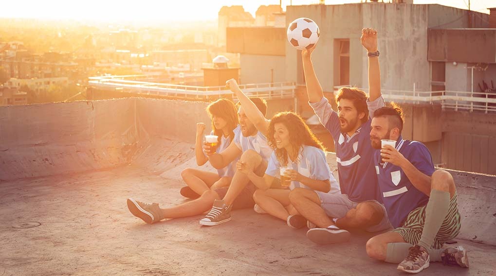 A group of friends sit on a rooftop with drinks, cheering. One person holds up a soccer ball, with city views at sunset.