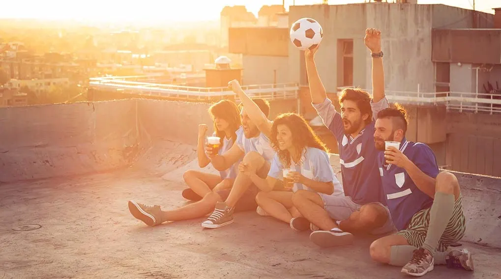 A group of friends sit on a rooftop with drinks, cheering. One person holds up a soccer ball, with city views at sunset.