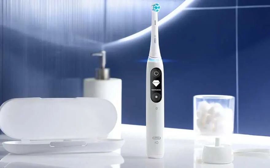 A white electric toothbrush stands on a bathroom counter beside a case, dispenser and glass against a blue backdrop.