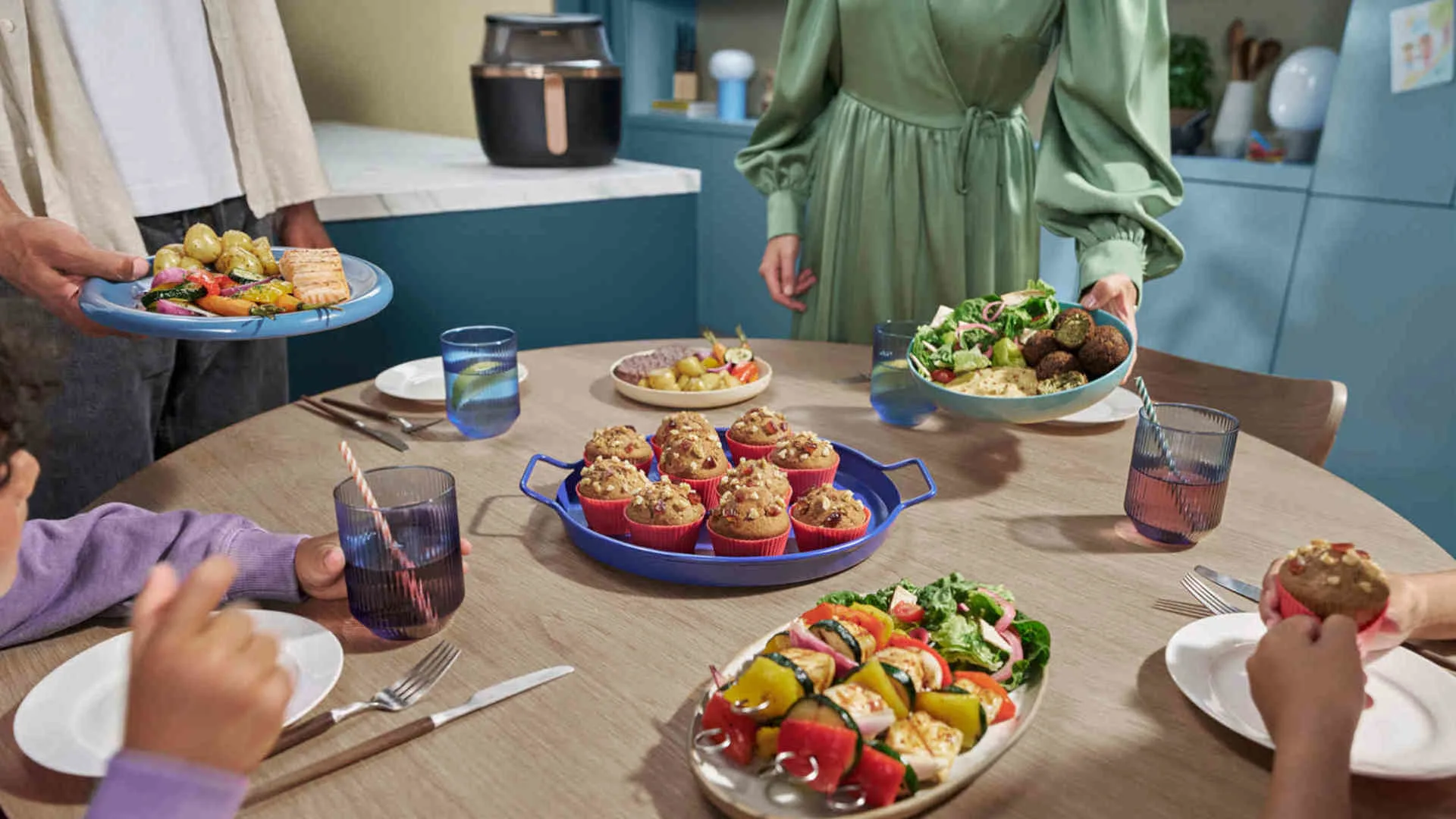 Ronde tafel met gerechten, salades, muffins en gegrilde groenten. Mensen delen maaltijden, met een heteluchtfriteuse op de achtergrond.