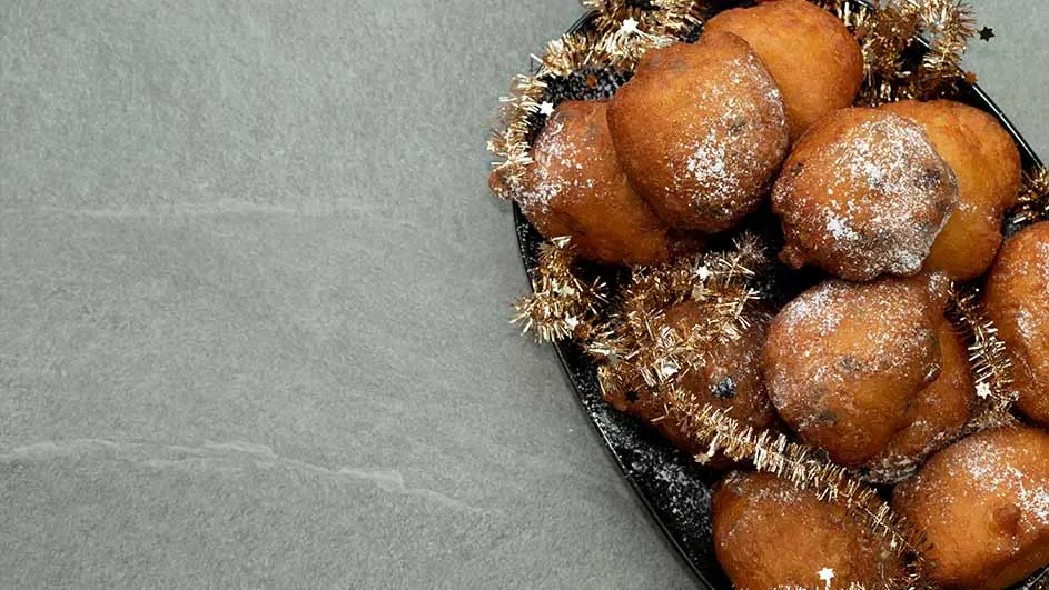 Oliebollen op een zwarte schaal met gouden slingers, bestrooid met poedersuiker, tegen een grijze achtergrond.