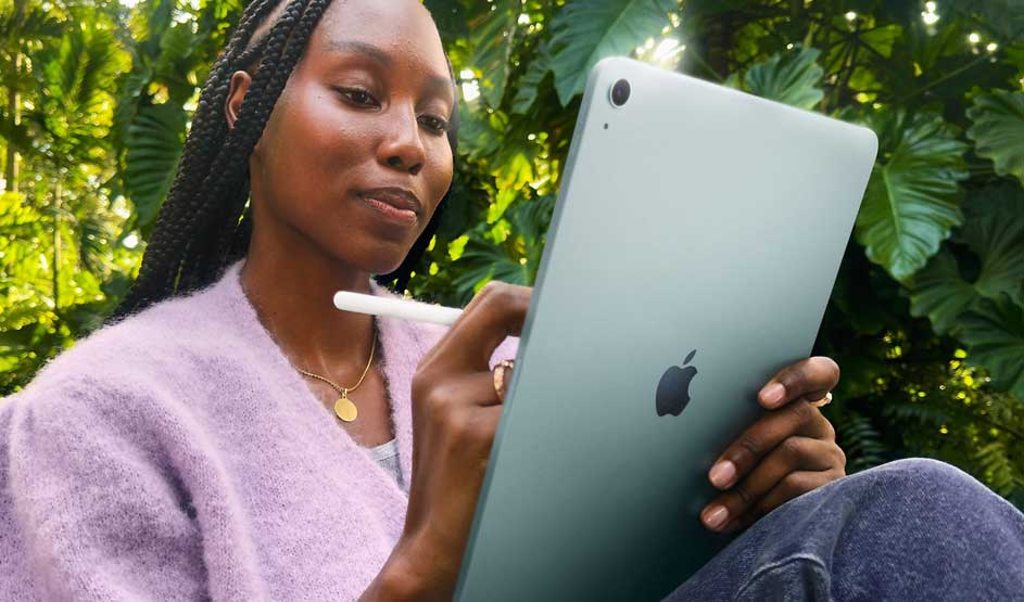 A woman sits outdoors among big leaves, using a stylus on her green iPad, looking relaxed yet focused on her creative work.