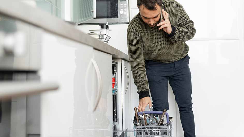 Een man in een groene trui laadt de vaatwasser in terwijl hij aan de telefoon is in een keuken.