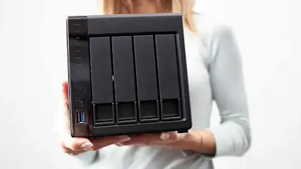 Een vrouw in een lichtgrijze trui houdt een zwarte NAS (Network Attached Storage) met vier schijfsloten voor zich vast. De achtergrond is egaal en lichtgrijs.