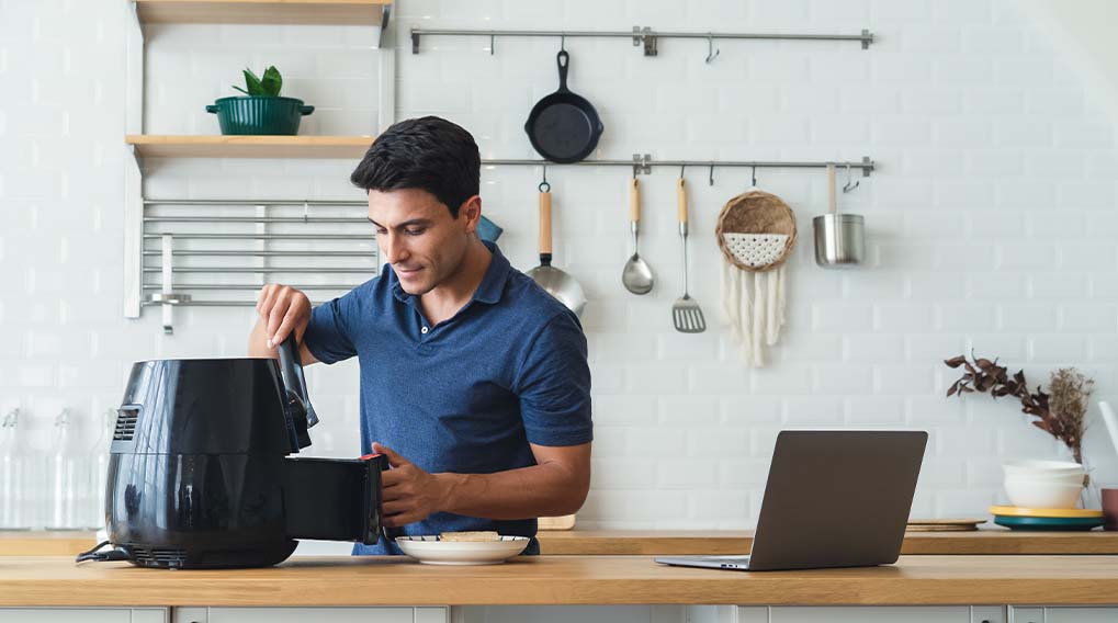 Een man in een blauw shirt bedient een zwarte airfryer in een keuken met keukengerei en een laptop.