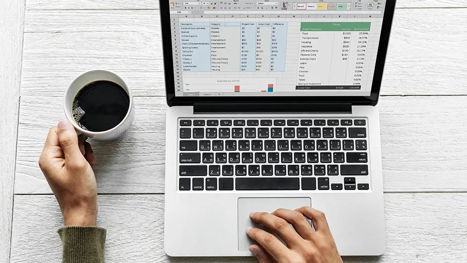 Laptop displays a spreadsheet on a white wooden desk, with a hand using the trackpad and another holding a coffee mug.