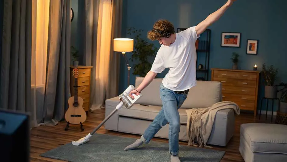 Een persoon stofzuigt een blauw tapijt in een woonkamer met een bank, gitaar en houten meubels.