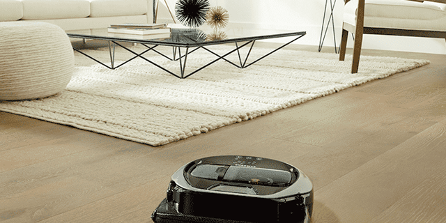 Een robotstofzuiger staat op een houten vloer, met een wit vloerkleed, een tafel en een bank in de achtergrond.