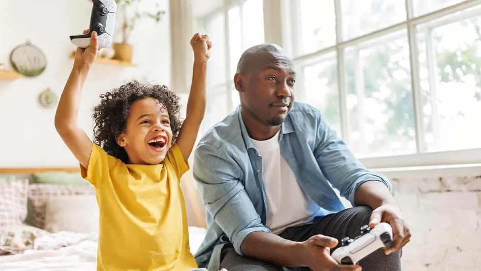 Een kind in een geel shirt juicht met een controller, terwijl een man met een controller kijkt, voor een raam.