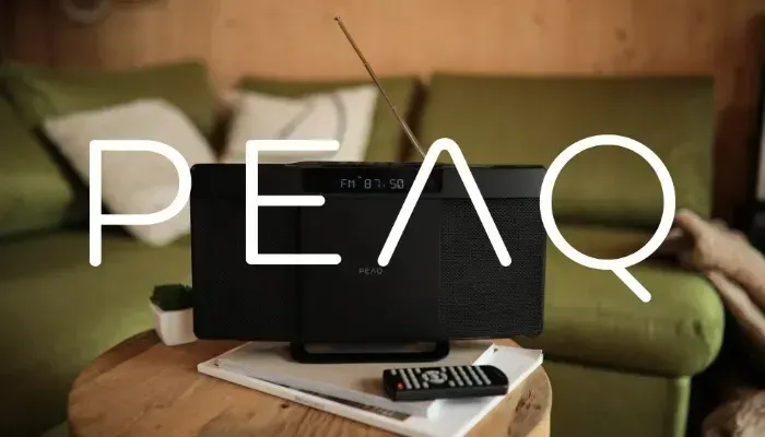 A black radio sits on a round wooden table with a remote and book. Behind it is a green couch with white pillows.