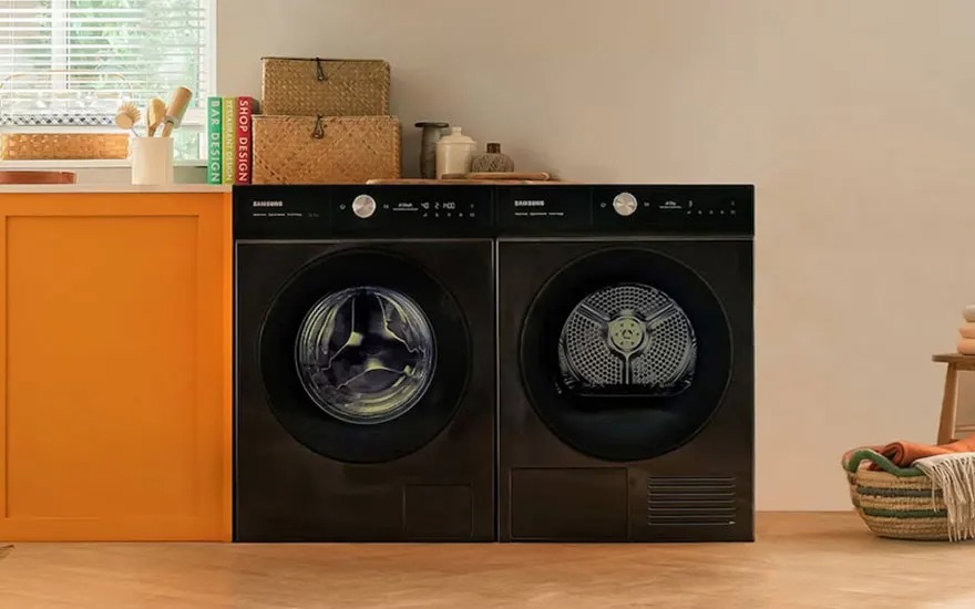 Een moderne wasruimte met zwarte Samsung was-droogcombinatie naast een oranje kast. Bovenop staan rieten manden en decoratie. Links een raam met jaloezieën, rechts een gevlochten wasmand op een houten vloer.