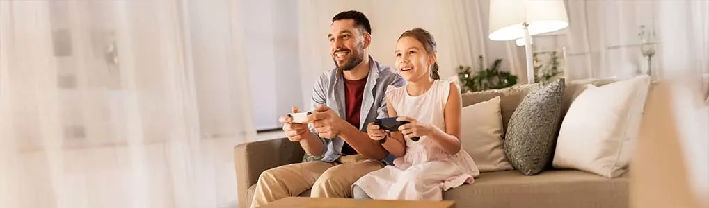Een glimlachende man en jong meisje zitten samen op een bank in een gezellige woonkamer, terwijl ze enthousiast gamen met elk een controller in de hand.
