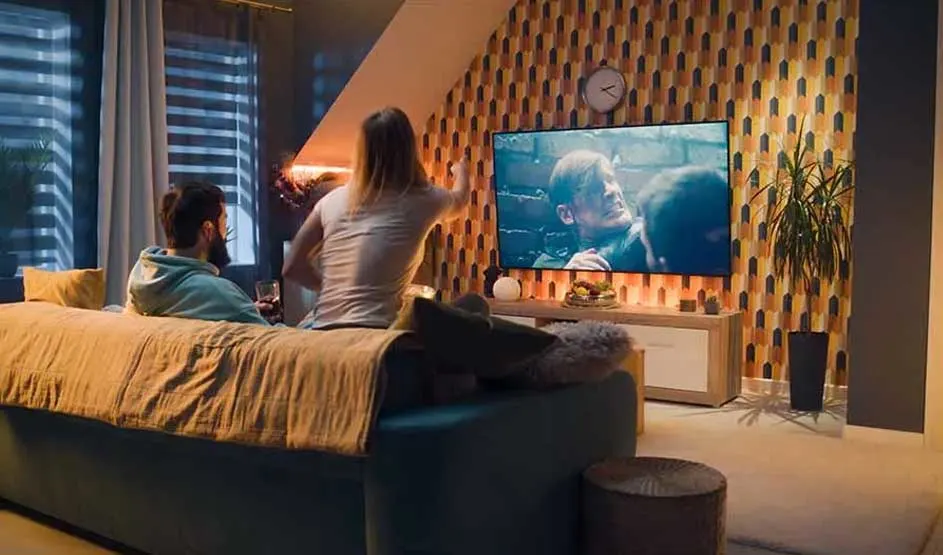 Two people watch television from bed in a warm, softly lit bedroom.