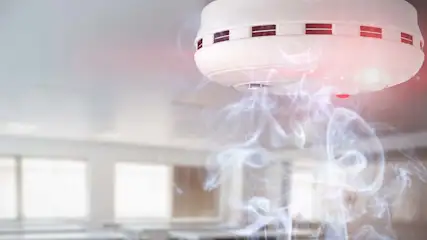 Witte rook onder een witte rookmelder aan een wit plafond. Ramen en tafels op de achtergrond.