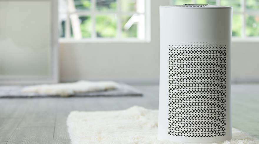 Een witte luchtreiniger met een driehoekig patroon staat op een witte vloerkleed, met een raam en spiegel op de achtergrond.