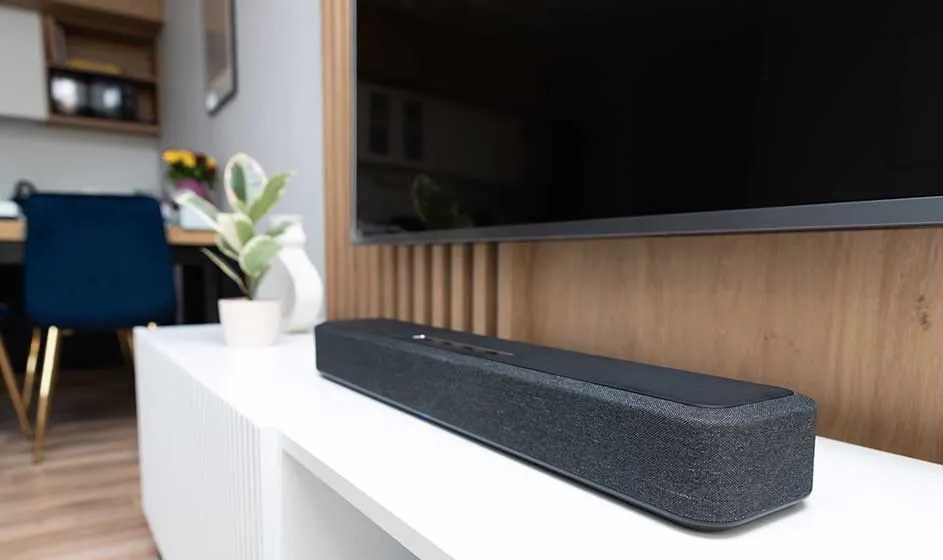 Een zwarte soundbar staat op een wit meubel onder een grote tv; hout, planten en een rustige kamer geven een moderne sfeer.